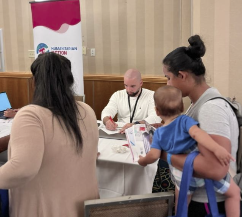 Joaquin Wilson working one-on-one with a participant at the SAMU First Response humanitarian intake desk, completing forms in front of a Humanitarian Action banner.